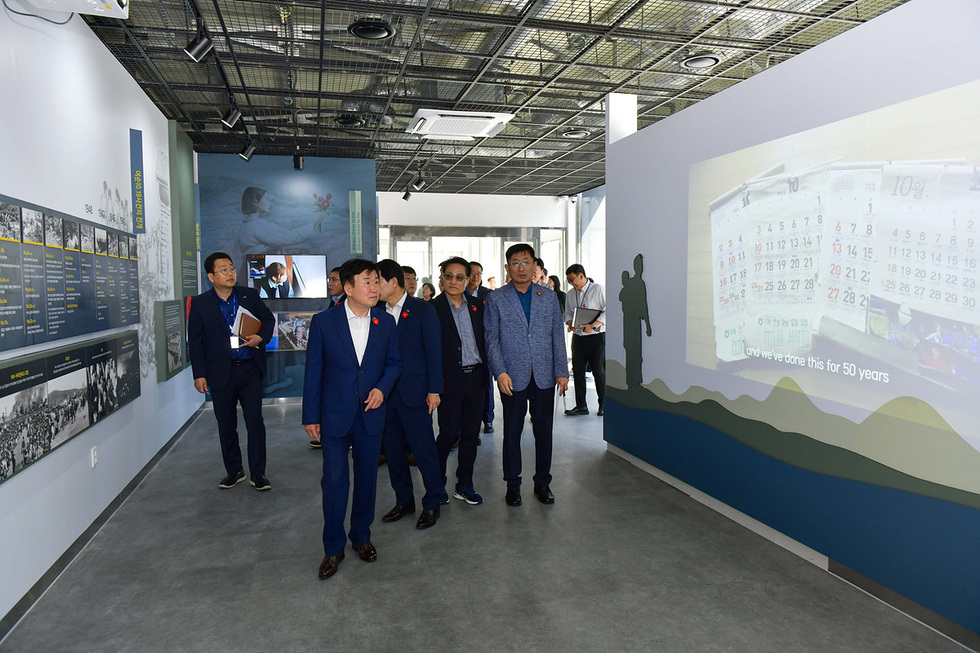 여러 명의 사람들이 전시관에서 전시물을 관람하며 대화하고 있는 장면이다. 벽에는 과거의 사진과 함께 50년 동안의 활동을 설명하는 내용이 담겨 있다.