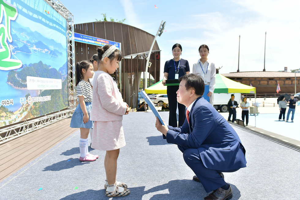 행사에서 한 남성이 어린 소녀에게 상장을 수여하고 있다. 배경에는 다른 어린이와 행사 관계자들이 보인다.