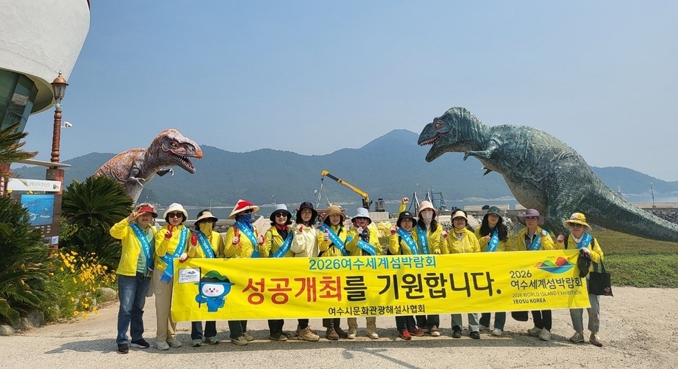여수에서 2026 세계섬박람회의 성공적인 개최를 기원하는 행사에서 참가자들이 현수막을 들고 포즈를 취하고 있다. 배경에는 공룡 모형이 보인다.