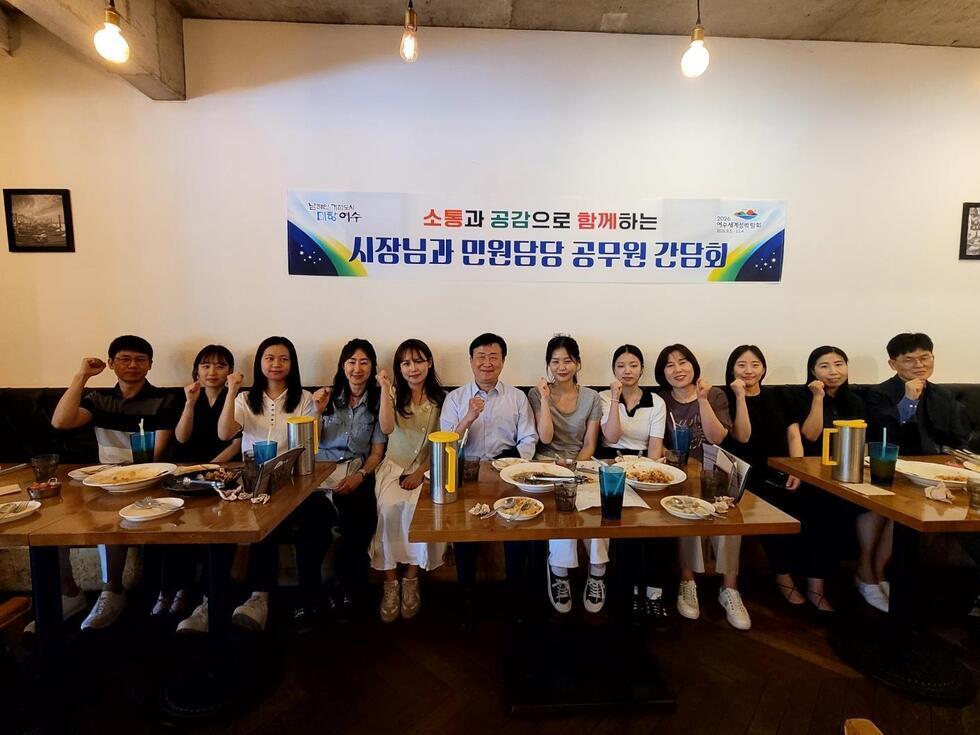 시청남과 민원당 공무원들이 간담회를 위해 모여 손을 흔들고 있는 모습이다. 테이블 위에는 음식과 음료가 놓여 있다.