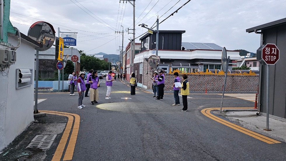 도로에서 보라색 조끼를 입은 사람들이 모여 있는 모습으로, 정지 표지판이 보인다.