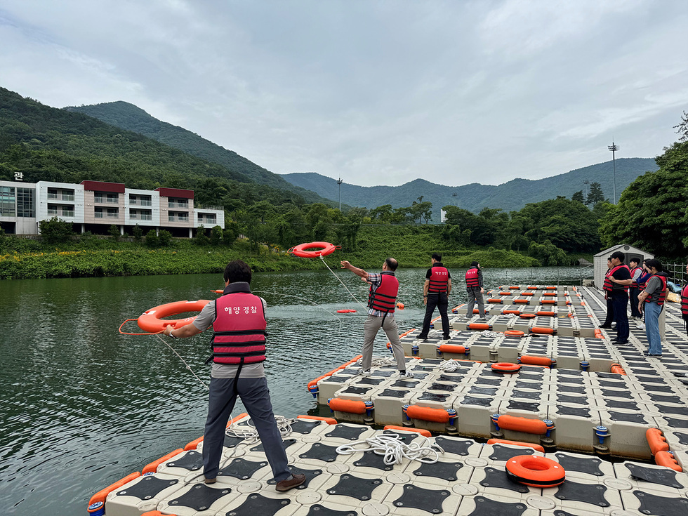 해양경찰 직원들이 수상에서 구명환을 던지는 훈련을 하고 있는 모습이다. 배경에는 산과 건물이 보인다.