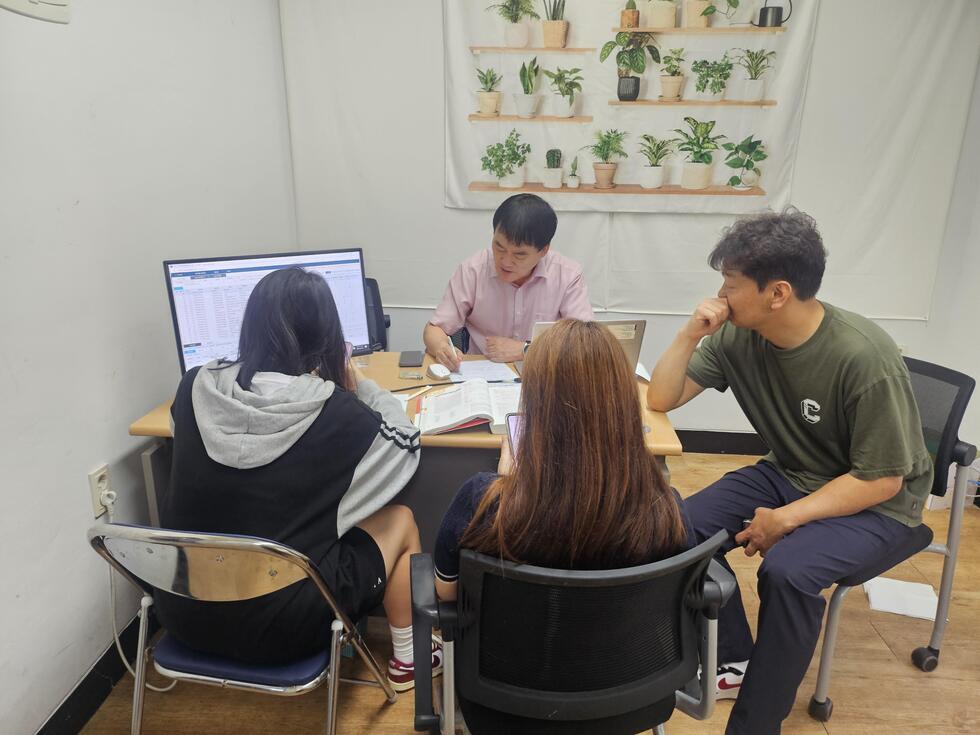 사무실에서 네 명이 모여 앉아 대화하고 있으며, 한 사람은 컴퓨터 화면을 보고 있다. 배경에는 식물이 장식된 벽이 보인다.