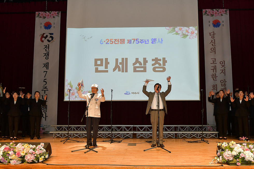 6 25전쟁 제75주년 행사에서 두 사람이 만세삼창을 하고 있으며, 뒤에는 참석자들이 손을 들고 있다.