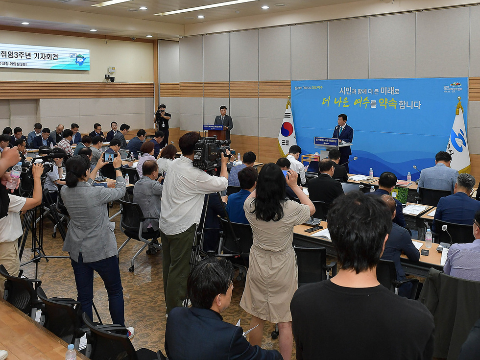 기자회견에서 연설하는 남성 앞에 기자들이 앉아 있으며, 배경에는 '시민과 함께 더 큰 미래로 더 나은 어식을 약속합니다'라는 문구가 적힌 현수막이 있다.