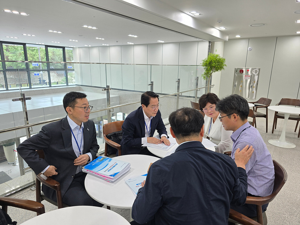 회의 중인 여러 명의 사람들이 테이블에 앉아 문서에 대해 논의하고 있는 장면이다. 배경에는 큰 창문과 식물이 보인다.