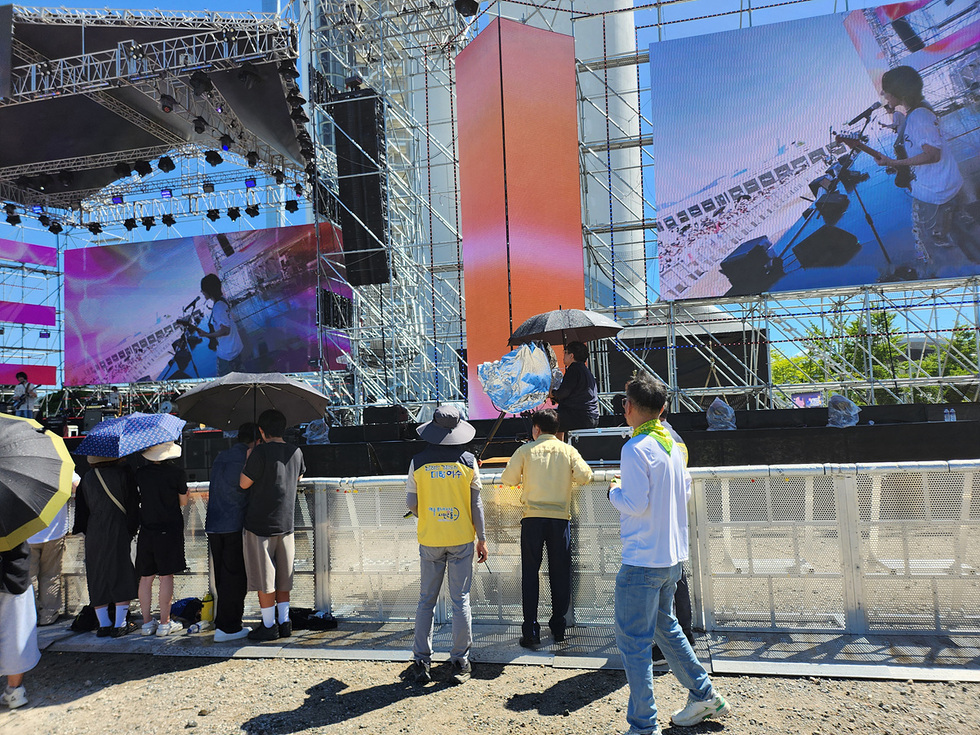 대형 무대 앞에서 여름 축제를 즐기는 관객들, 폭염 속에서 일부는 양산과 모자로 햇빛을 가리고 공연을 관람하는 모습