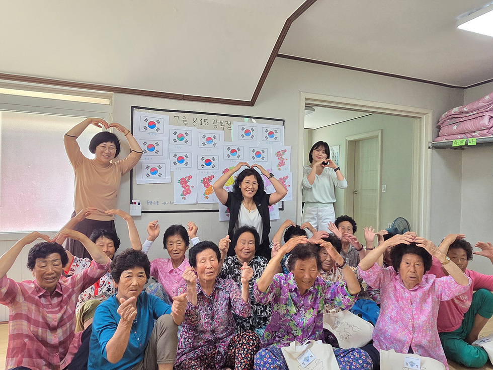 르신들과 봉사단원이 함께 광복절을 기념하며 단체 사진을 찍는 모습. 모두 손으로 하트 모양을 만들며 밝게 웃고 있음.