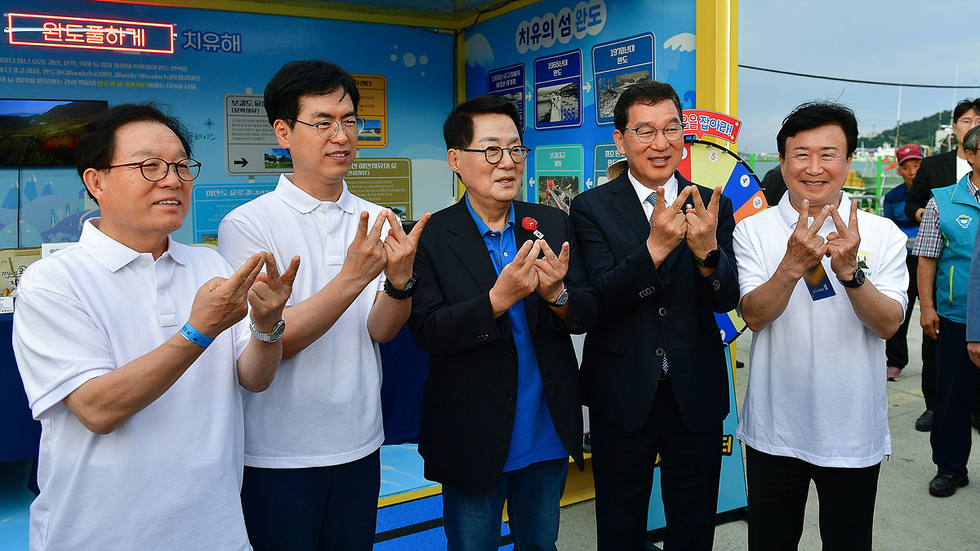 다섯 명의 남성이 흰색 티셔츠를 입고 손가락으로 V자를 만들며 포즈를 취하고 있다. 배경에는 치유와 관련된 정보가 담긴 패널이 있다.