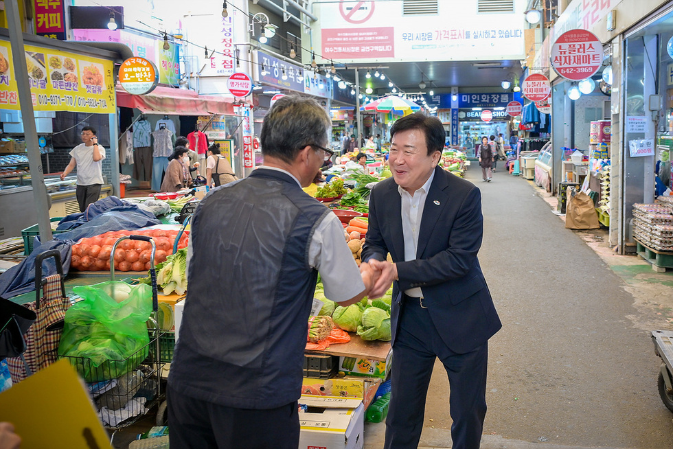 시장 내에서 두 남성이 악수하며 대화하고 있는 모습으로, 주변에는 다양한 채소와 과일이 진열되어 있다.