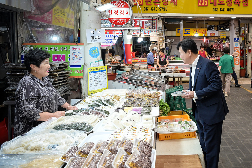 시장 한쪽에서 여성 상인이 떡을 판매하고 있으며, 남성이 떡을 살펴보고 있다. 배경에는 다른 상인들이 활동하는 모습이 보인다.