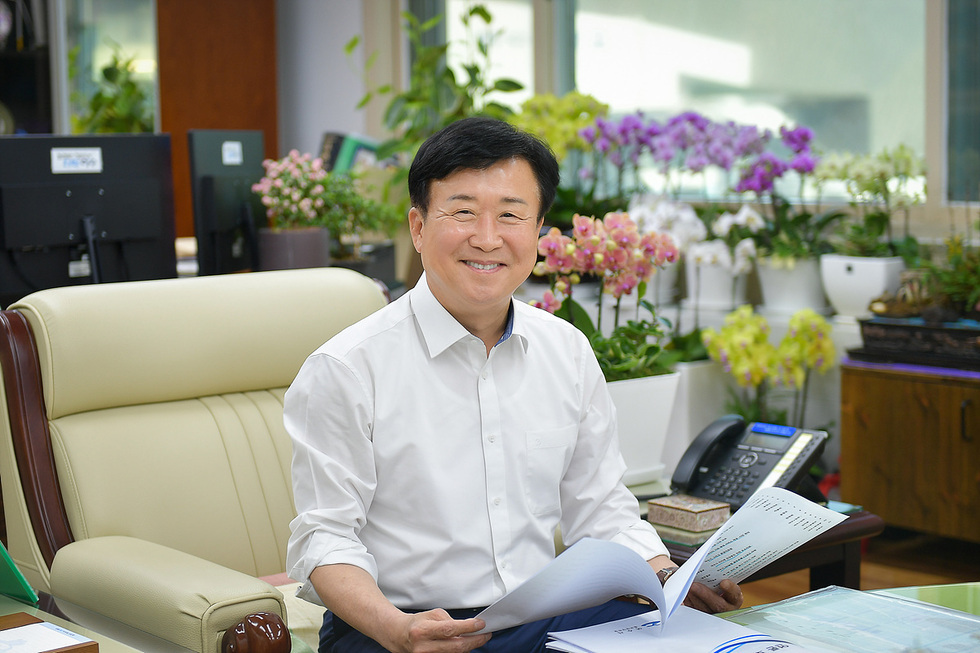 사무실에서 서류를 들고 웃고 있는 남성이 앉아 있다. 배경에는 여러 종류의 꽃이 있는 화분이 놓여 있다.