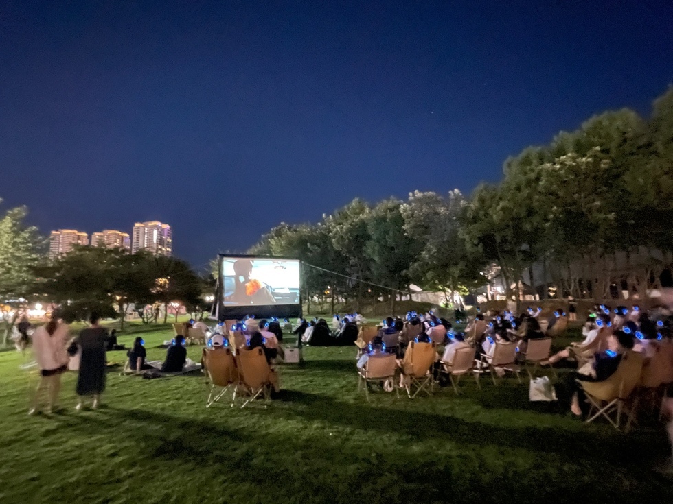 야외에서 사람들이 헤드폰을 착용하고 대형 스크린에서 영화를 관람하는 장면이다. 주변에는 나무와 건물이 보인다.