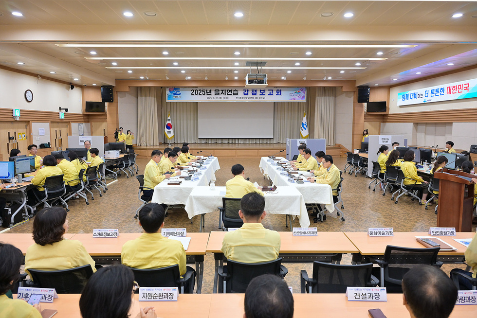 2025년 을지연습 강평보고회가 진행되고 있는 모습으로, 참석자들이 노란색 옷을 입고 회의 중이다. 배경에는 대한민국 국기가 걸려 있고, 회의 테이블에는 여러 부서명이 적힌 이름표가 놓여 있다.