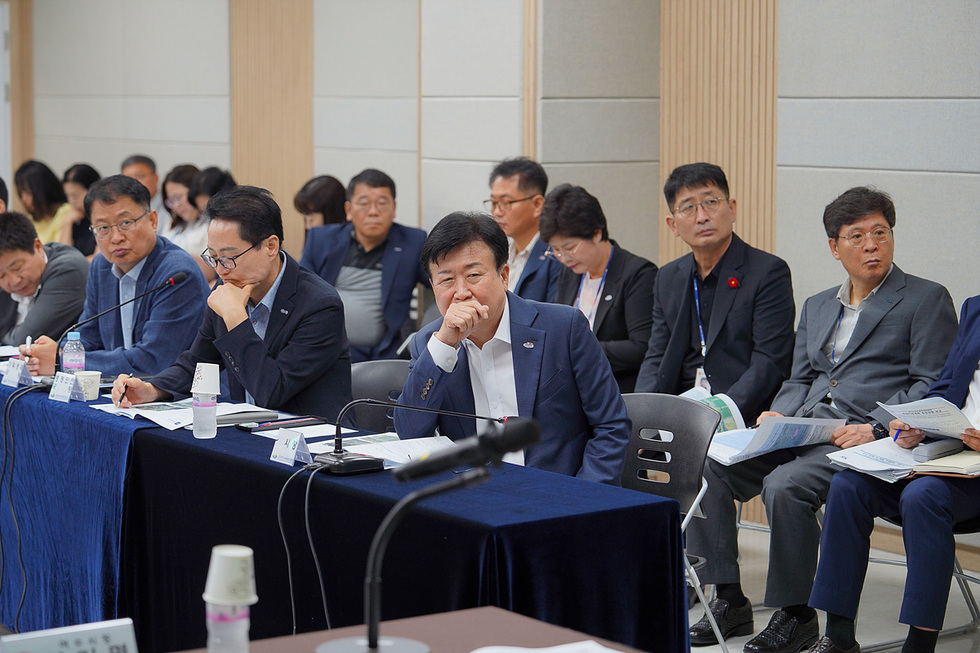 회의 중인 여러 명의 사람들이 앉아 있는 모습으로, 중간에 한 남성이 손으로 얼굴을 가리고 생각에 잠겨 있다.