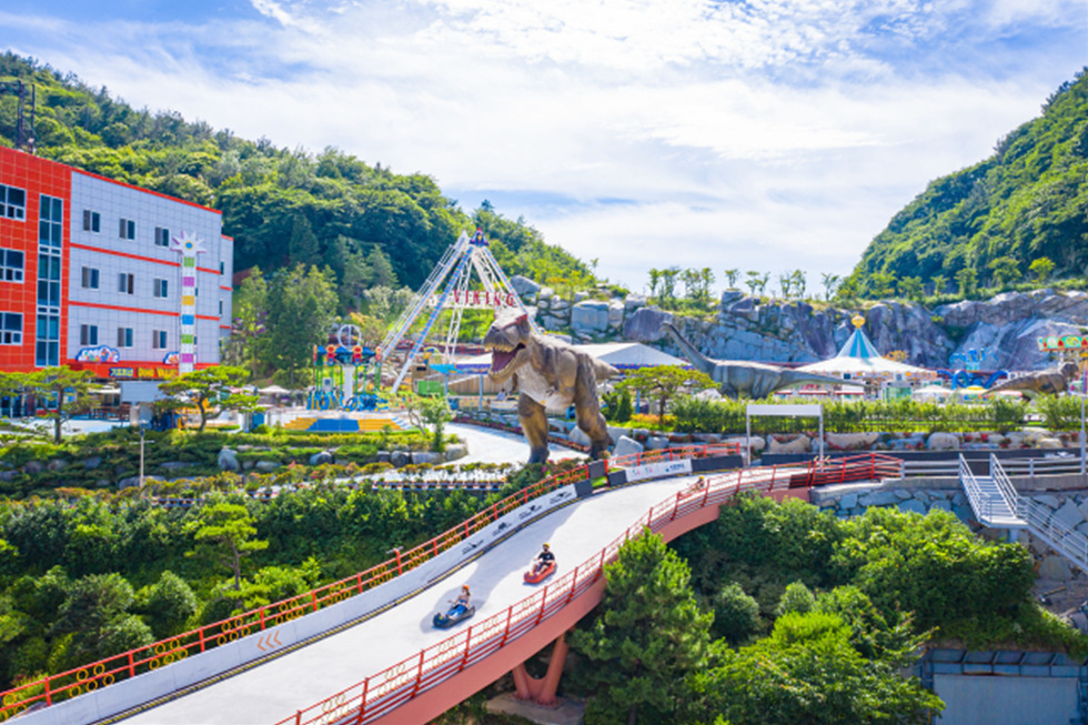 놀이공원에서 공룡 모형이 있는 풍경과 함께 사람들이 카트에 타고 있는 모습