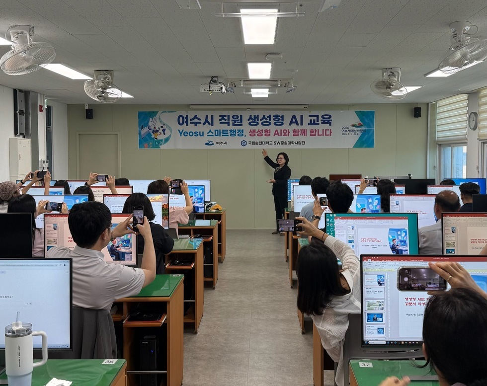 여수시에서 진행되는 직업형 AI 교육 현장으로, 강사가 화면을 가리키며 설명하고 있으며, 교육생들이 스마트폰으로 촬영하고 있다.