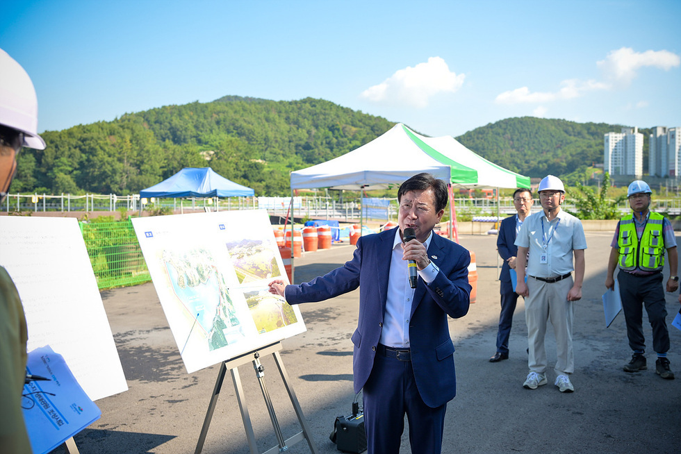 한 남성이 마이크를 들고 설명하며 지도 앞에서 포즈를 취하고 있다. 뒤에는 여러 명의 사람들이 서 있고, 텐트와 공사 장비가 보인다.