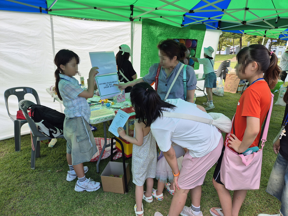 아이들이 부스에서 도움을 요청하며 활동하고 있는 모습으로, 테이블에는 다양한 물품이 놓여 있다.