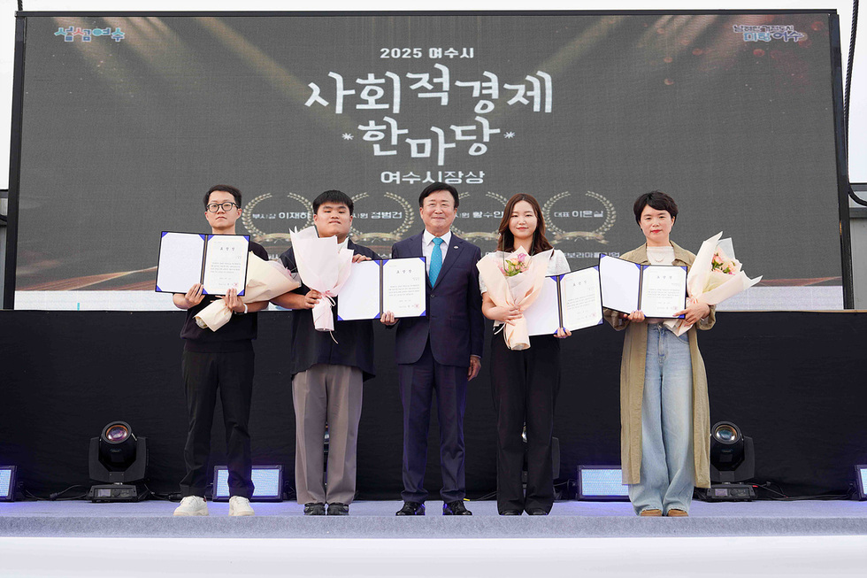 정기명 시장이 무대 위에서 연설하는 가운데 객석에 앉은 시민들과 관계자들이 경청하고 있으며, 뒤편에는 사회적경제 기업 부스가 줄지어 설치된 사회적경제 한마당 행사장 전경.