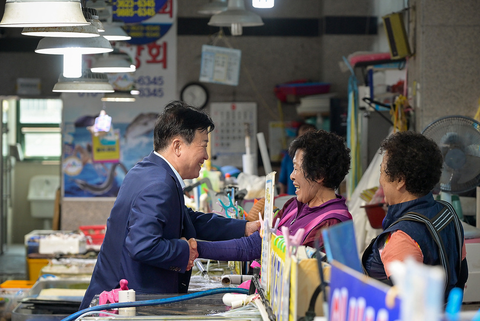 정기명 여수시장이 전통시장 내 수산물 판매 상인과 반갑게 악수하며 대화하는 모습. 주변에 수산물이 진열되어 있고 상인들이 환하게 웃고 있다.