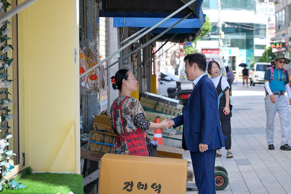 여수시장이 길가 점포 앞에서 여성 상인과 악수하며 인사를 나누는 장면. 상인은 건어물 상자 옆에 서 있으며, 주변으로 시장 풍경과 시민들이 보인다.