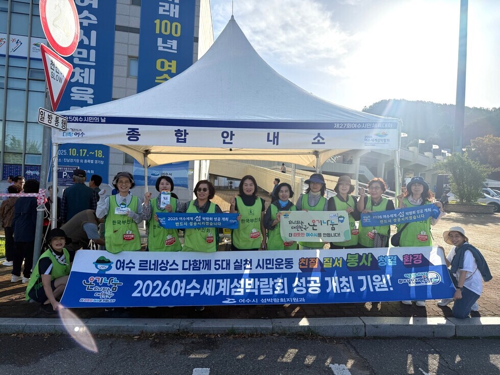 여수시민의 날 행사에서 자원봉사자들이 2026여수세계섬박람회 홍보를 위해 여수 르네상스 다함께 5대 실천 시민운동 칠전 질서 봉사 청결 환경 2026년 여수 세계섬박람회 성공 개최 기원 현수막을 들고 있는 모습