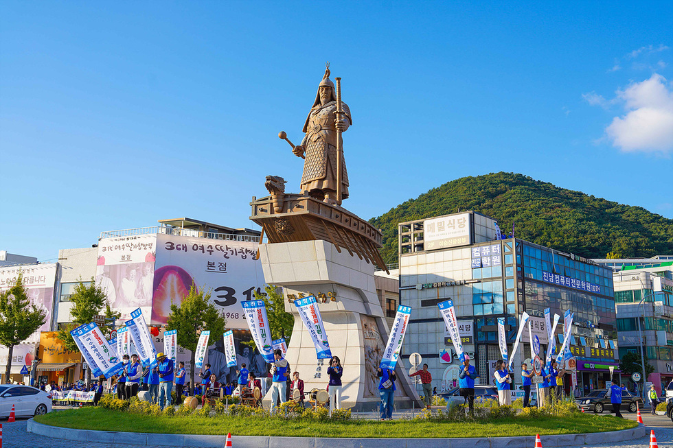 여수 중앙동로터리에서 시민들이 친절과 청결을 주제로 한 캠페인에 참여하고 있는 모습으로, 깃발을 들고 구호를 외치고 있다.