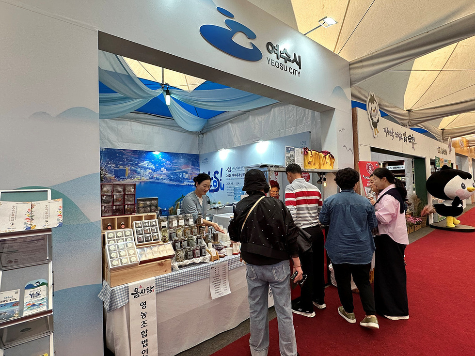 여수시 홍보관에서 사람들이 지역 농특산물을 구매하고 있는 모습으로, 부스에는 다양한 제품이 진열되어 있다.