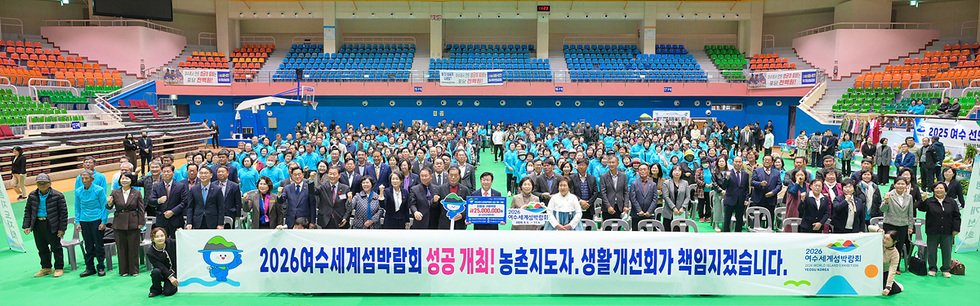 진남실내체육관에서 수백 명의 참석자가 단체로 기념사진을 촬영하고 있으며, 앞쪽에는 ‘2026여수세계섬박람회 성공 개최! 농촌지도자·생활개선회가 책임지겠습니다.’라고 적힌 긴 현수막이 펼쳐져 있다.