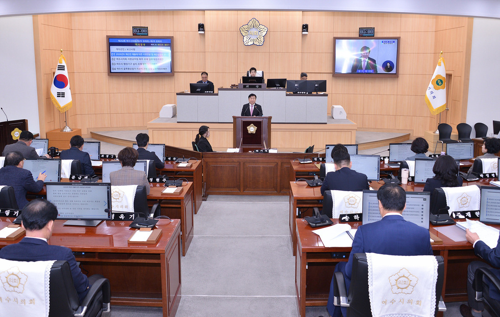 여수시의회 본회의장에서 여러 의원들이 좌석에 앉아 회의를 진행하고 있으며, 중앙 연단에서 정기명 여수시장이 발언하고 있다. 좌측에는 대한민국 국기, 우측에는 여수시의회 깃발이 비치되어 있다. 앞 전광판에는 회의 안건이 표시되어 있다.
