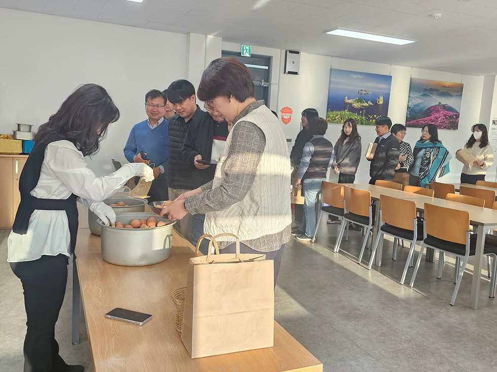 회의실 식탁 앞에서 직원들이 줄을 서서 큰 냄비에 담긴 구운계란을 받아가는 모습. 직원 한 명이 앞쪽에서 종이봉투를 들고 계란을 나눠주고 있으며 뒤쪽으로 여러 직원들이 차례로 줄을 서 있다