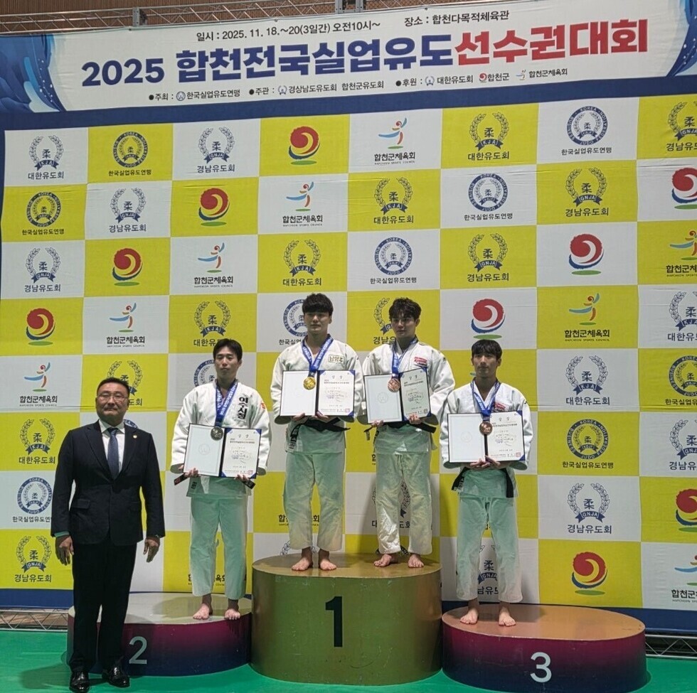 합천 전국 실업유도선수권대회 시상식 무대에서 남자 선수 4명이 포디움 위에 올라 금·은·동메달을 걸고 상장을 들고 있는 모습. 왼쪽에는 정장 차림의 관계자가 함께 촬영하고 있다. 배경에는 대회 명칭이 적힌 노란색·흰색 패턴의 포토존이 설치되어 있다.