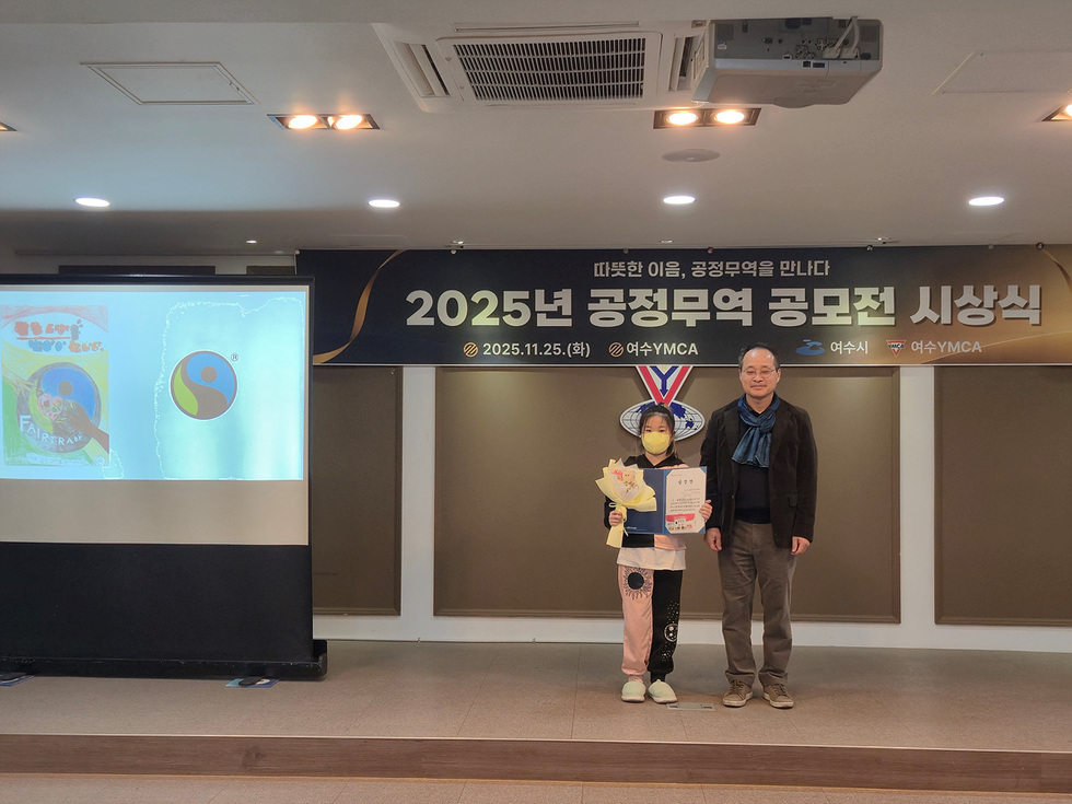 공정무역 공모전 시상식 무대에서  수상자가 상장과 꽃다발을 들고 기념 촬영을 하고 있다. 뒤편에는 ‘2025년 공정무역 공모전 시상식’이라고 적힌 현수막이 걸려 있고, 무대 왼쪽 화면에 공모전 관련 이미지가 비쳐지고 있다.