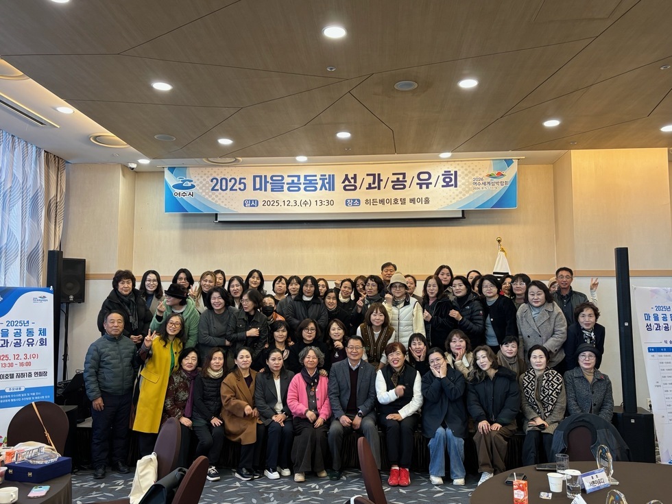 2025 여수시 마을공동체 성과공유회 행사장에서 히든베이호텔 베이홀을 배경으로 참가자 40여 명이 무대 앞에 모여 단체 기념촬영을 하고 있으며, 상단 현수막에는 ‘2025 마을공동체 성과공유회 / 일시 2025.12.3.(수) 13:30 / 장소 히든베이호텔 베이홀’이라는 문구가 적혀 있고 양쪽에는 행사 안내 배너가 세워져 있다.