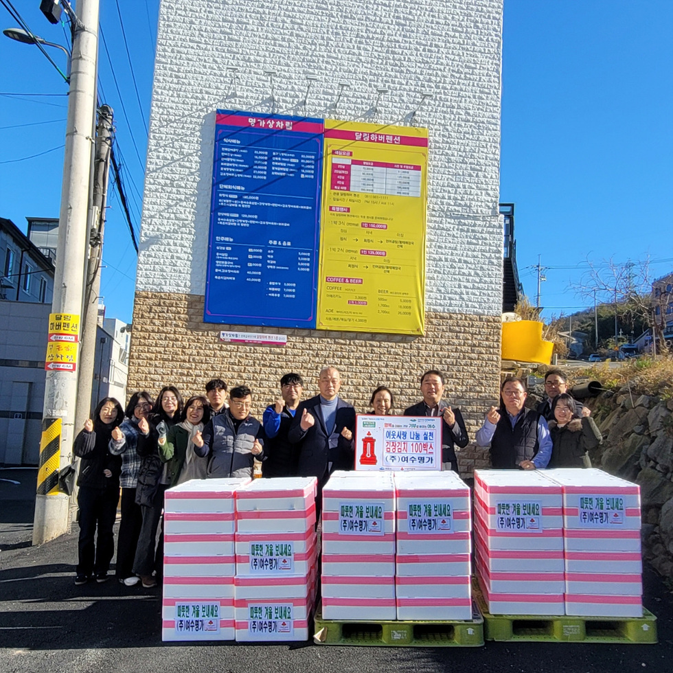여수명가 건물 앞에서 여러 관계자들이 김장 김치 상자 100박스와 함께 나눔 전달식을 진행하고 있으며, 참여자들이 박스 앞에 일렬로 서서 손하트를 하고 있고 상자에는 ‘따뜻한 겨울 보내세요’ 문구가 붙어 있으며 안내판에는 ‘여수사랑 나눔 실천 김장김치 100박스 기증’ 내용이 적혀 있다.