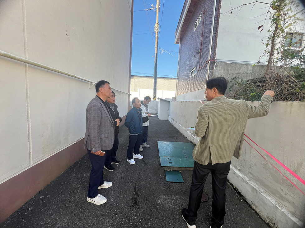 여수시 공동주택 재난 대비 취약시설 점검을 위해 관계자들이 주택 사이 골목에서 옹벽과 배수시설 등을 살펴보고 있다. 점검 담당자가 벽면을 가리키며 설명하고 있으며, 다른 참석자들은 현장을 관찰하며 메모를 하고 있다.