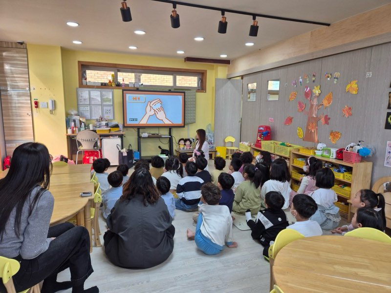 어린이들이 교실에서 화면을 바라보며 수업을 듣고 있다. 화면에는 손을 사용하는 방법을 설명하는 그림이 나타나 있다.