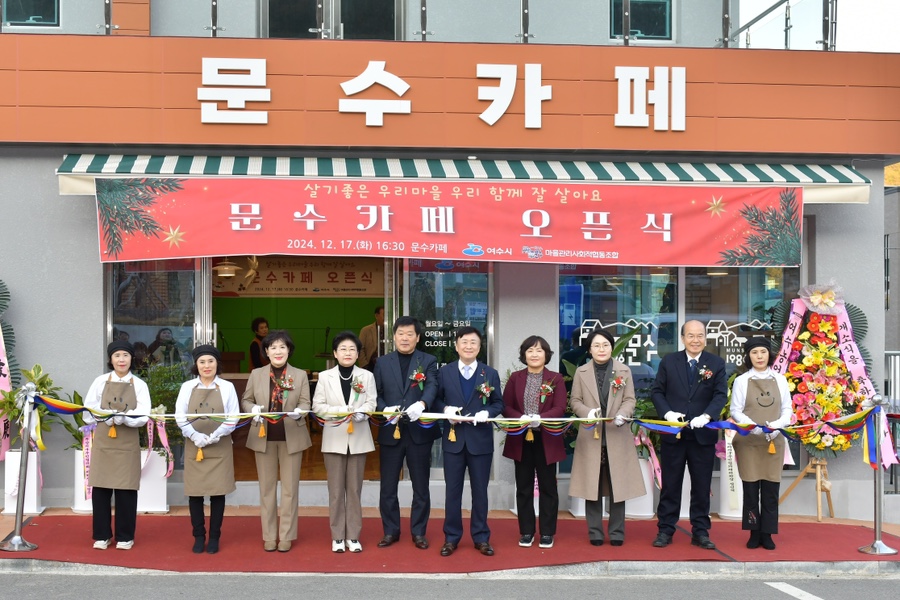 문수카페 개업식에서 여러 사람들이 리본을 자르고 있다. 카페 외부에는 개업을 알리는 현수막과 꽃다발이 놓여 있다.