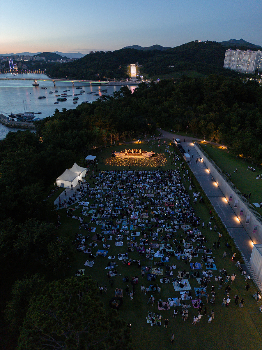 해변가에서 많은 사람들이 모여 야외 공연을 관람하고 있는 모습이다. 주변에는 바다와 산이 보이며, 저녁 하늘이 물들어 있다.