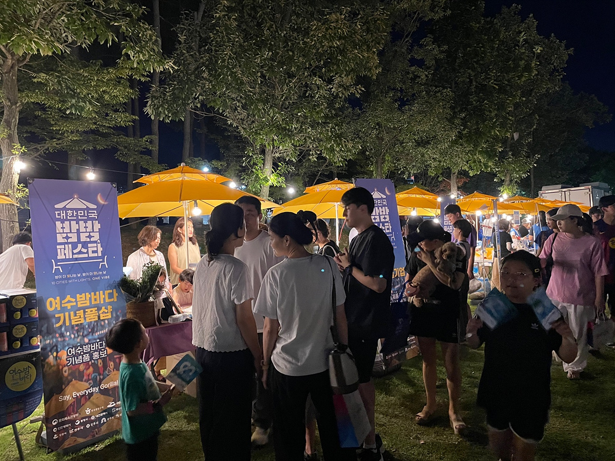 여수밤바다 기념품샵에서 사람들이 모여 있는 모습으로, 노란 파라솔 아래에서 다양한 기념품을 판매하고 있다.