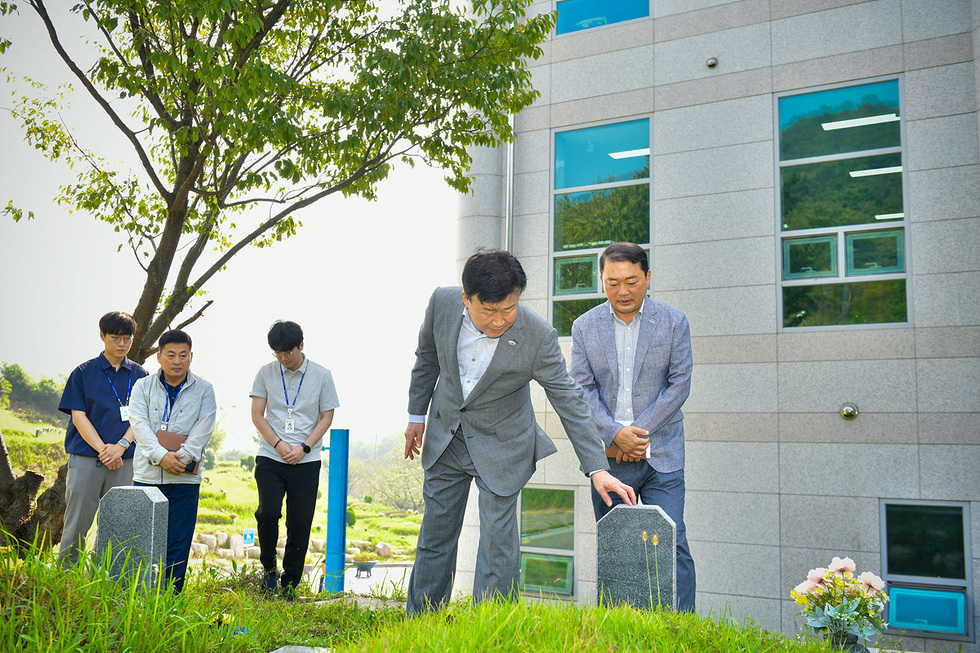 한 남성이 묘비에 손을 대고 있으며, 다른 세 명의 남성이 그를 바라보고 있는 장면이다. 배경에는 건물과 나무가 보인다.