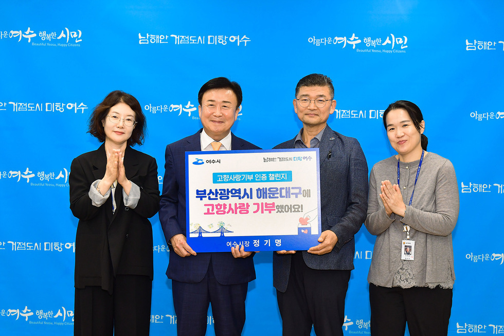 여수시청에서 고향사랑기부 인증 챌린지를 기념하는 행사로, 여수시장 정기명이 부산 해운대구에 기부한 내용을 담은 현수막을 들고 있다. 참석자들이 함께 기념사진을 찍고 있다.