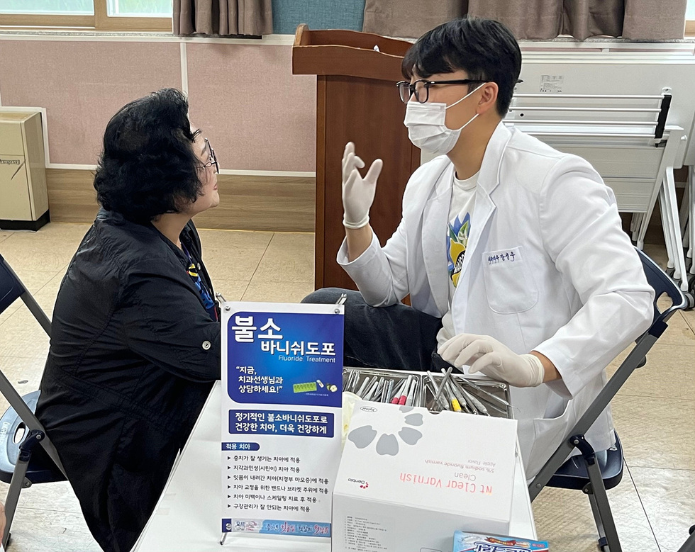 치과의사가 환자와 상담하고 있는 장면으로, 불소 바니쉬 치료에 대한 안내가 적힌 현수막이 보인다.