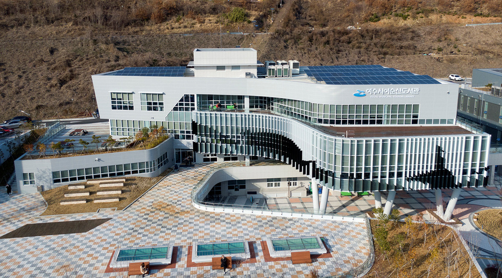 여수시립도서관의 외관이 보이며, 건물에는 태양광 패널이 설치되어 있다. 주변에는 정원과 벤치가 배치되어 있다.