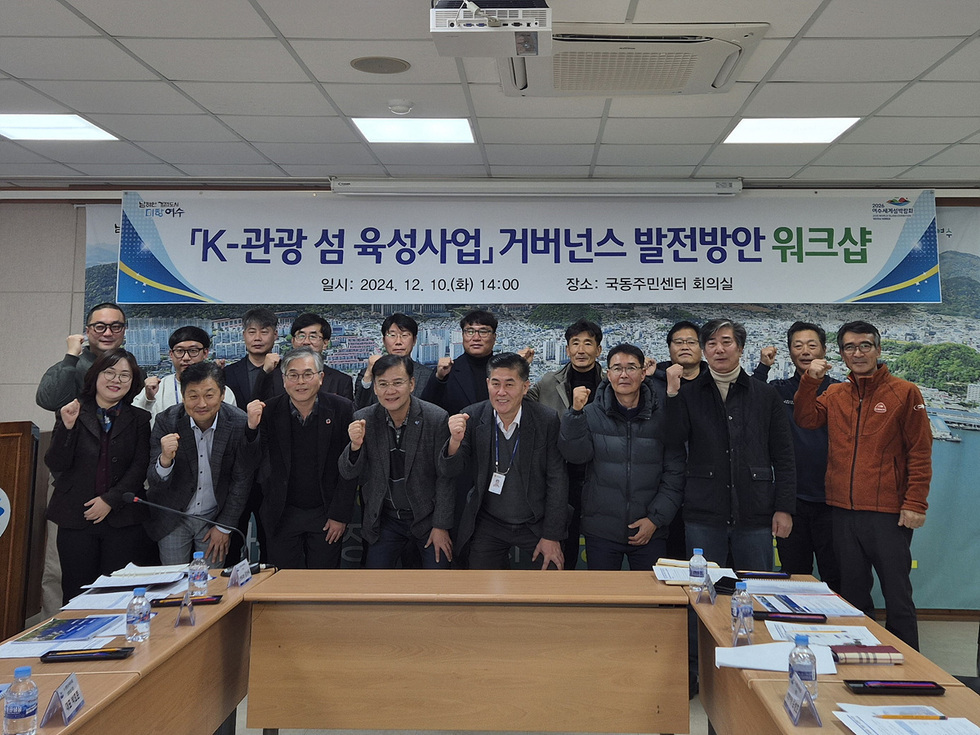 K-관광 섬 육성사업과 관련된 워크샵에 참석한 사람들이 주먹을 쥐고 포즈를 취하고 있다. 배경에는 행사 제목과 일시, 장소가 적힌 현수막이 걸려 있다.