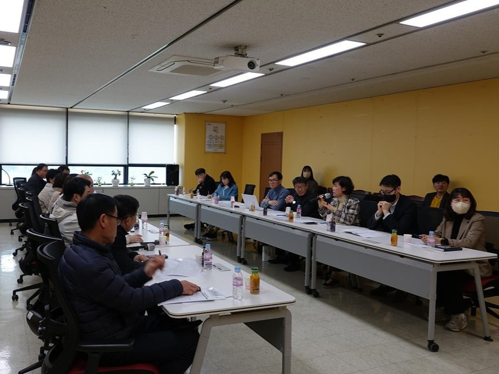 회의실에서 여러 사람이 테이블에 앉아 회의 중인 모습이다. 참석자들은 문서와 음료수를 앞에 두고 의견을 나누고 있다.