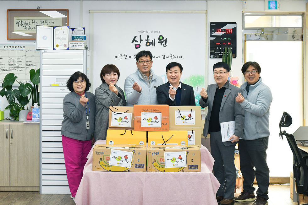 사무실에서 여섯 명의 사람들이 상자를 앞에 두고 손가락 하트를 만들며 기념사진을 찍고 있다.