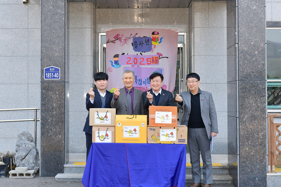네 명의 남성이 상자 앞에서 손가락 하트를 만들며 포즈를 취하고 있다. 배경에는 '을사년 2025福'이라는 문구가 적힌 현수막이 걸려 있다.