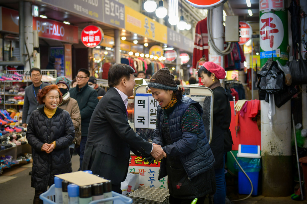 시장 한복판에서 한 남성이 여성과 악수하며 대화하고 있다. 주변에는 여러 사람들이 쇼핑을 하고 있는 모습이 보인다.